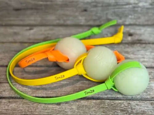 Trois balles tug phosphorescentes pour chiens avec poignées en Biothane orange, jaune et verte de différentes longueurs, posées sur un fond de bois.